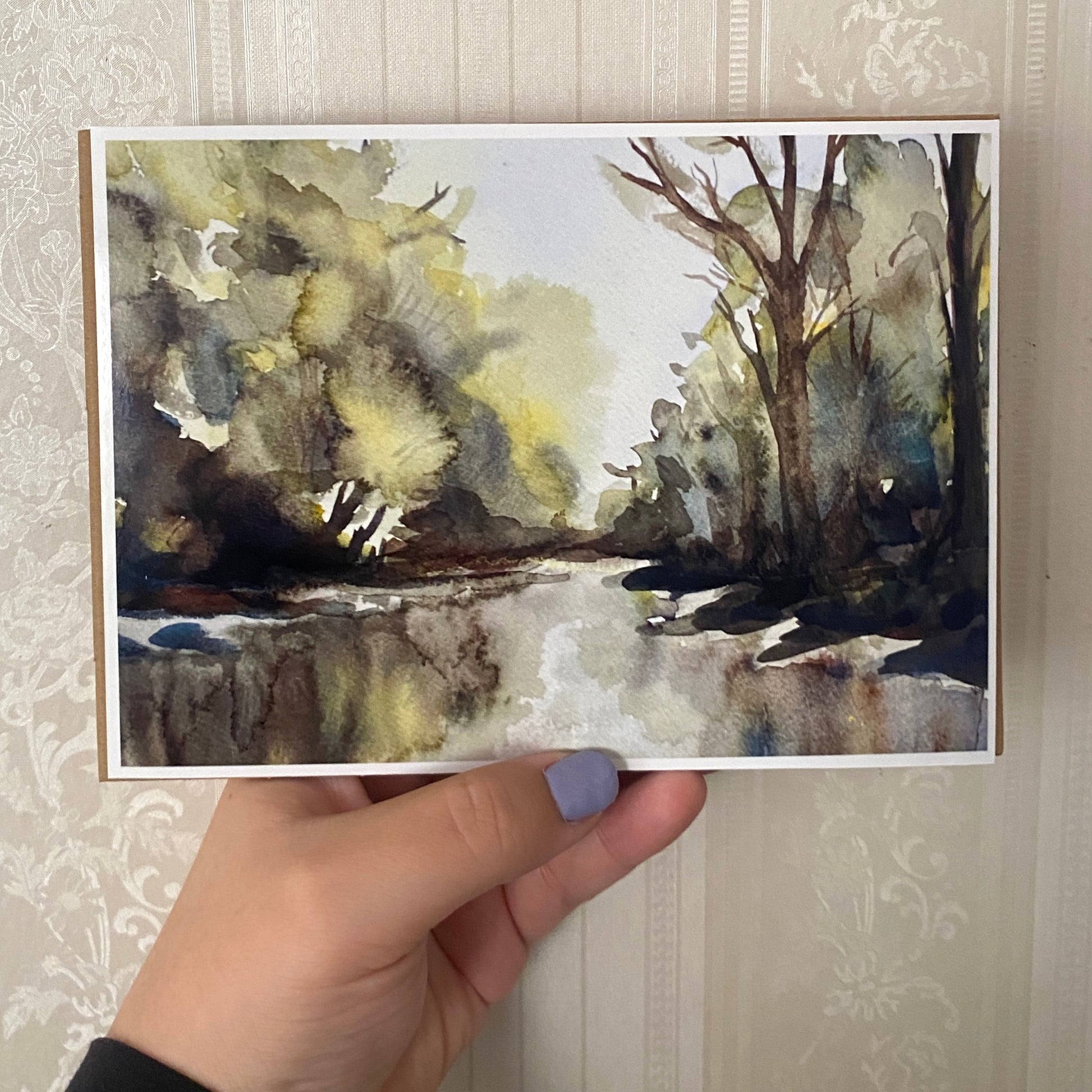 Hand holding a watercolor painting of a forest scene with trees and a stream.